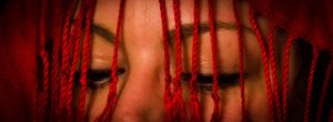Ausschnitt der Augen einer Frau über deren Gesicht rote Fäden hängen.